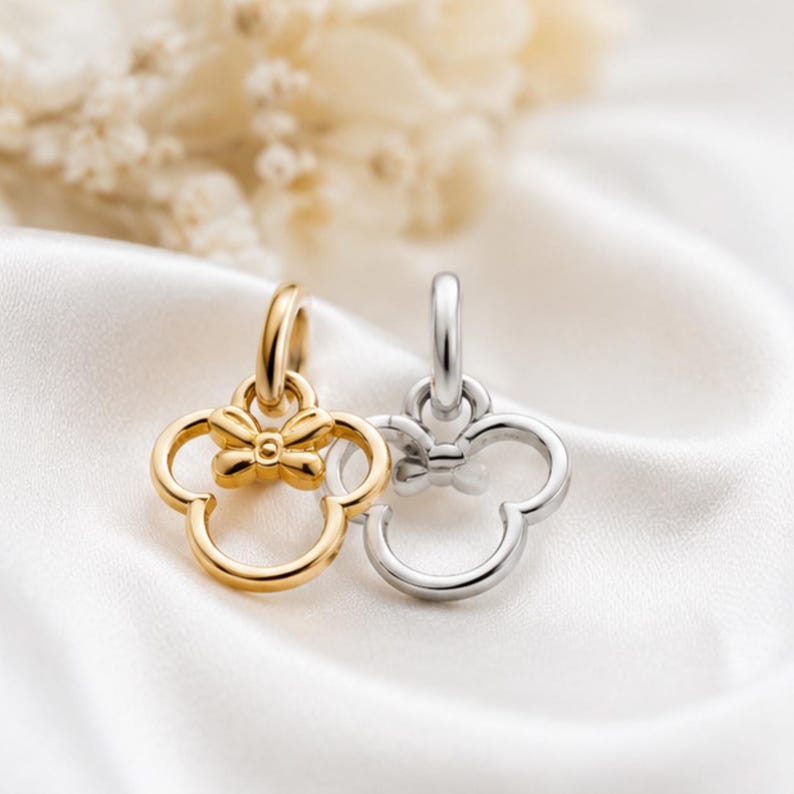 Puede incluir: Dos colgantes de metal, uno dorado y otro plateado, cada uno con forma de cabeza estilizada con orejas redondeadas y un peque&ntilde;o lazo en la parte superior. Cada colgante cuenta con una anilla circular para sujetarlo a una cadena o collar.