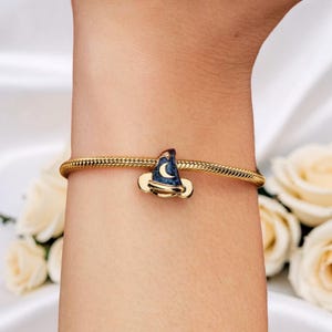 May include: A gold-colored bracelet with a charm shaped like a wizard's hat. The hat is dark blue with a crescent moon and stars design. The charm is accented with gold-colored metal. The bracelet is worn on a wrist.