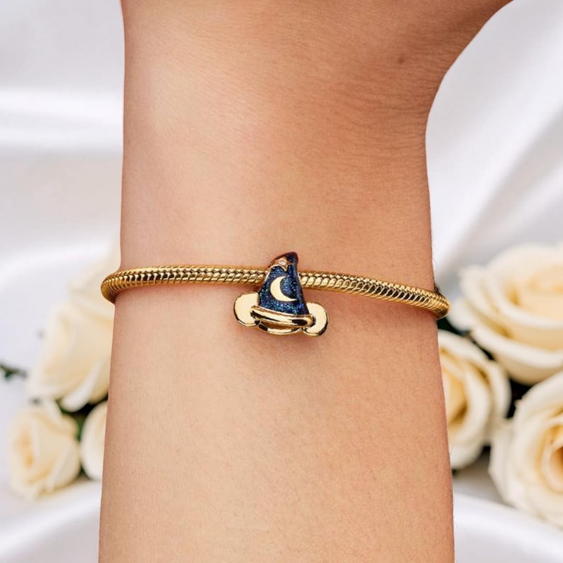 May include: A gold-colored bracelet with a charm shaped like a wizard's hat. The hat is dark blue with a crescent moon and stars design. The charm is accented with gold-colored metal. The bracelet is worn on a wrist.