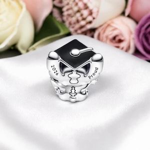 May include: A silver charm with a black graduation cap design. The charm has the year "2026" and the word "Proud" engraved on it. The charm is on a white surface with blurred flowers in the background.
