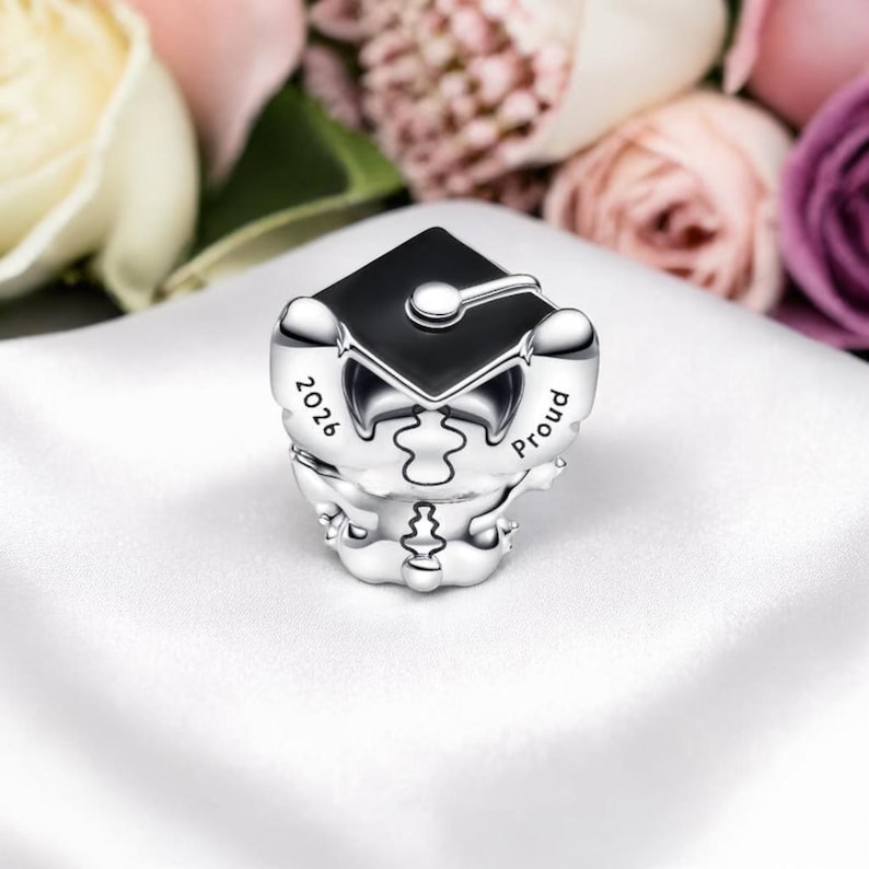May include: A silver charm with a black graduation cap design. The charm has the year "2026" and the word "Proud" engraved on it. The charm is on a white surface with blurred flowers in the background.