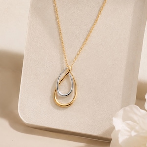 May include: A two-tone pendant necklace featuring a gold-coloured chain and a teardrop-shaped charm. The charm consists of a larger gold-coloured teardrop loop intertwined with a smaller silver-coloured teardrop loop, creating a layered design.