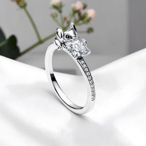 May include: A silver-toned ring featuring a sculpted character head with large ears and a square-cut clear gemstone setting. The band is accented with a row of small, round, clear stones along the side, creating a detailed and polished appearance.