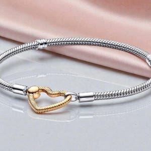May include: A silver bracelet with a gold heart-shaped clasp. The bracelet has a textured, snake-like design. The gold heart clasp is in the centre of the bracelet. The background is a soft pink fabric.