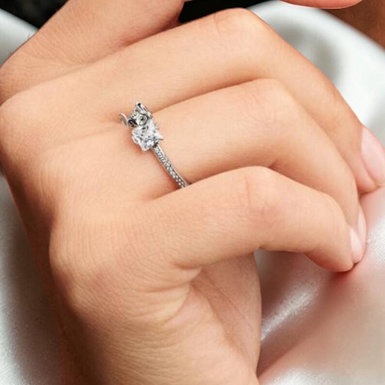 May include: A silver-toned ring featuring a square-cut clear gemstone set in a prong setting. The band is slender and decorated with a series of small, embedded clear stones that extend along the visible portion of the metal ring shank.