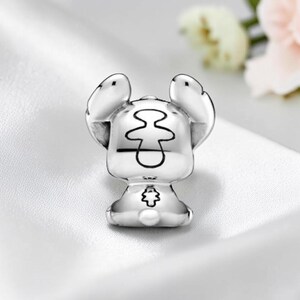 May include: A silver-toned metal charm shaped like a stylized creature with large, upright ears. The back of the charm features a central etched outline resembling a figure and a small, detailed floral-like emblem near the base of the rounded body.