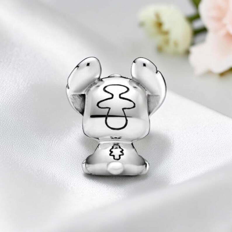 May include: A silver-toned metal charm shaped like a stylized creature with large, upright ears. The back of the charm features a central etched outline resembling a figure and a small, detailed floral-like emblem near the base of the rounded body.