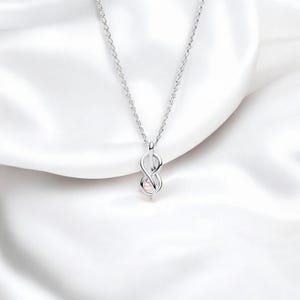 May include: A silver-colored necklace with an infinity symbol pendant. The pendant has a small, light pink pearl at its center. The necklace hangs on a white, silky background.