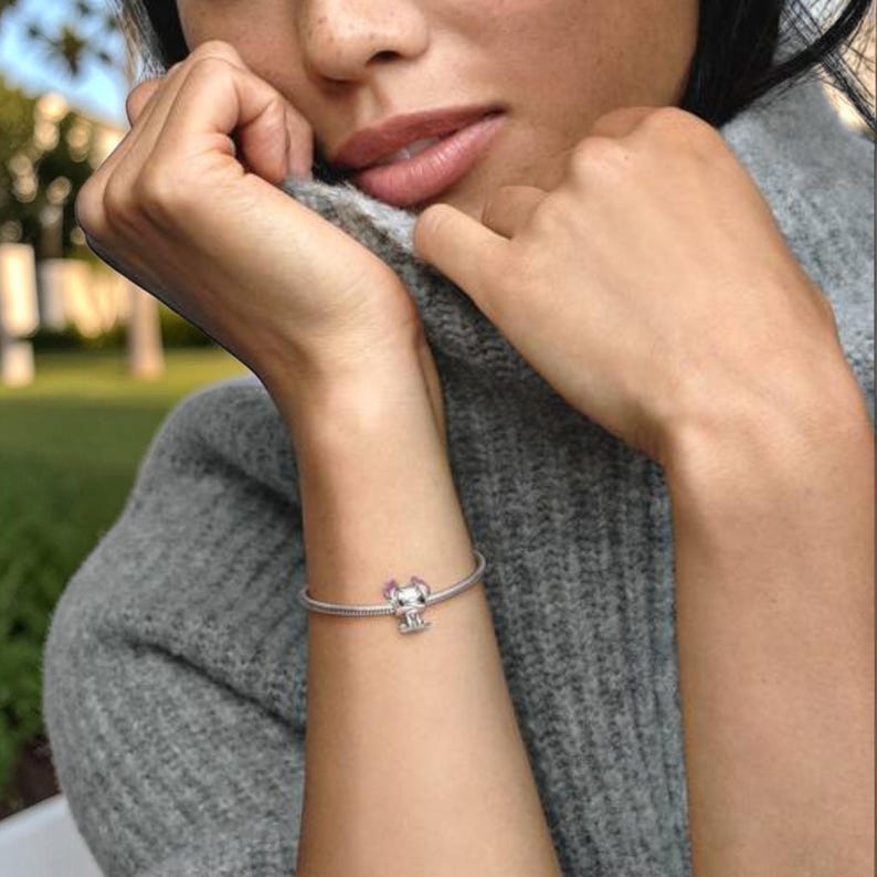 May include: A silver snake chain bracelet featuring a single charm shaped like a small character with pink accents. The bracelet is worn around the wrist, showcasing the metallic finish of the chain and the detailed design of the central charm.