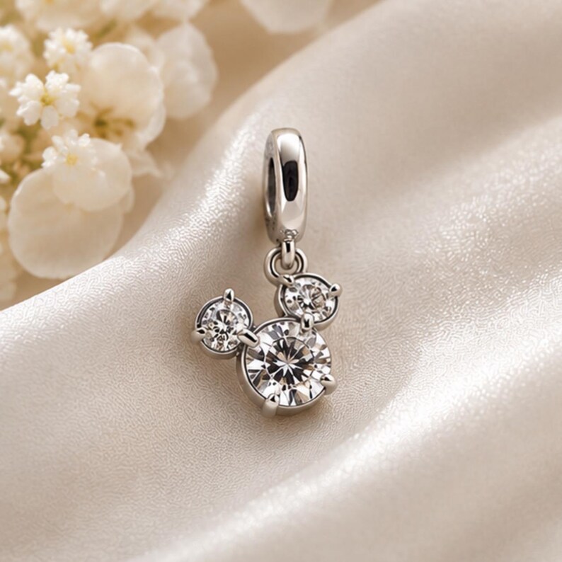 May include: A silver-toned charm featuring three clear, round-cut stones arranged in a cluster that resembles a circular silhouette. The charm includes a polished metal bail at the top for attachment, with each stone secured by small metal prongs.