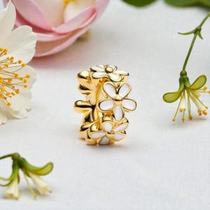 May include: A gold-colored charm with white enamel daisy flower designs. The charm is a floral-themed jewelry piece, with a bright and shiny appearance. The daisies are arranged in a stacked pattern, creating a three-dimensional effect.