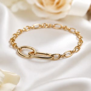 Könnte beinhalten: Ein goldfarbenes Kettenarmband mit einer Reihe gleichmäßiger ovaler Glieder, die mit einem größeren, länglichen ovalen Glied verbunden sind. Das Metall hat eine polierte, reflektierende Oberfläche, die ein glattes, kreisförmiges Design bildet.