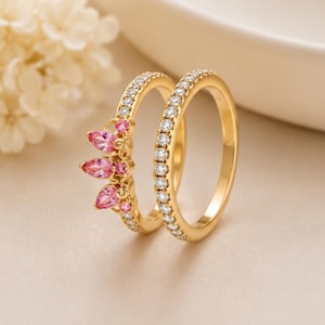 Pode incluir: Um conjunto de dois anéis dourados. Um anel apresenta uma fila de pedras transparentes, enquanto o outro inclui um design em forma de coroa com três pedras cor-de-rosa em forma de pera e pedras cor-de-rosa redondas menores em metal dourado.