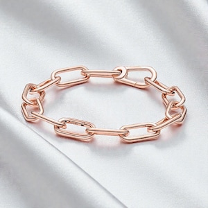 May include: Rose gold chain link bracelet. The bracelet features a series of interconnected oval links, creating a modern and elegant design. The metal has a warm, pinkish hue, and the clasp is integrated into the design. The bracelet is displayed on a white satin surface.