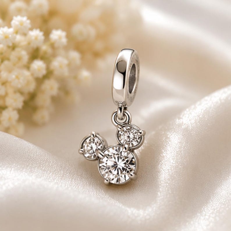 May include: A silver-toned metal charm featuring three round, clear, faceted stones arranged in a cluster resembling a mouse head silhouette. The charm includes a polished metal bail at the top for attachment to a bracelet or necklace chain.