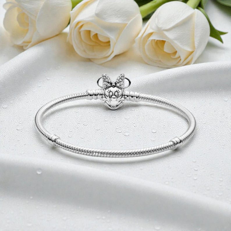 May include: A silver snake chain bracelet featuring a clasp shaped like a cartoon mouse head with a bow. The clasp is embellished with small clear stones. The bracelet has two fixed stations along the chain to hold charms in place.