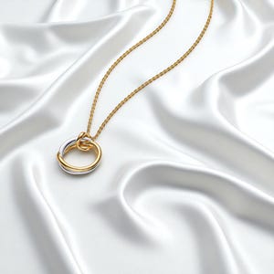 May include: A gold and silver necklace with a knot pendant. The pendant has two interlocking rings, one gold and one silver, forming a knot. The chain is a fine gold colour, set against a white satin background.