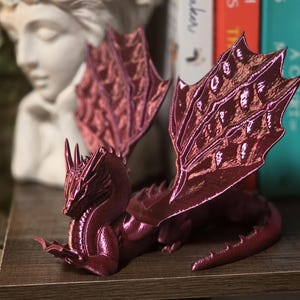 Może przedstawiać: Maroon figurka smoka ze szczegółowymi skrzydłami i teksturowanym korpusem. Smok leży na drewnianej półce. Skrzydła mają odblaskową, opalizującą jakość. Figurka to dekoracyjny przedmiot o tematyce fantasy.