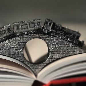 Op de afbeelding: Een gedetailleerde, grijze metalen trein- en bruggenboekensteun. De trein staat op een stenen brug, rustend op de pagina's van een open boek. De pagina's van het boek zijn wit met een rode rug.