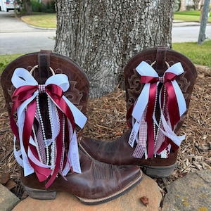 May include: A pair of brown leather cowboy boots adorned with decorative bows featuring maroon, white, and checkered ribbons, along with white beaded strands. The ribbons on the right boot clearly display the text AGGIES in white lettering.