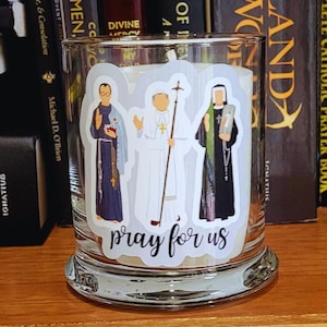 Może przedstawiać: Szklany słoik na świecę z trzema postaciami religijnymi na naklejkach. Postacie to mężczyzna w niebieskiej szacie, mężczyzna w białej szacie i kobieta w czarnej i zielonej szacie. Tekst "módl się za nas" jest napisany pod postaciami.