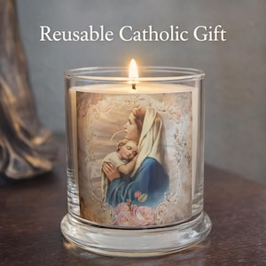 May include: A burning candle in a clear glass jar, featuring an image of Mary and baby Jesus. The candle sits on a clear base, with the text "Reusable Catholic Gift" above. The image is on a dark wooden surface.