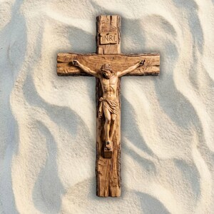 May include: A wooden crucifix with a carved figure of Jesus Christ. The cross is made of rough-hewn wood, with the letters INRI at the top. The figure is detailed, with a draped cloth around the waist. The background is a textured, light-colored surface.