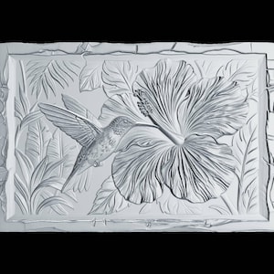 May include: A rectangular white wall plaque featuring a high-relief carving of a hummingbird hovering near a large hibiscus flower. The background displays detailed tropical foliage, and the entire piece has a textured, stone-like border.