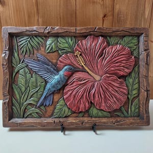 May include: A rectangular wooden wall plaque featuring a relief carving of a blue hummingbird with a red throat hovering near a large, textured red hibiscus flower. The scene is surrounded by green tropical leaves and a rustic, dark brown frame.