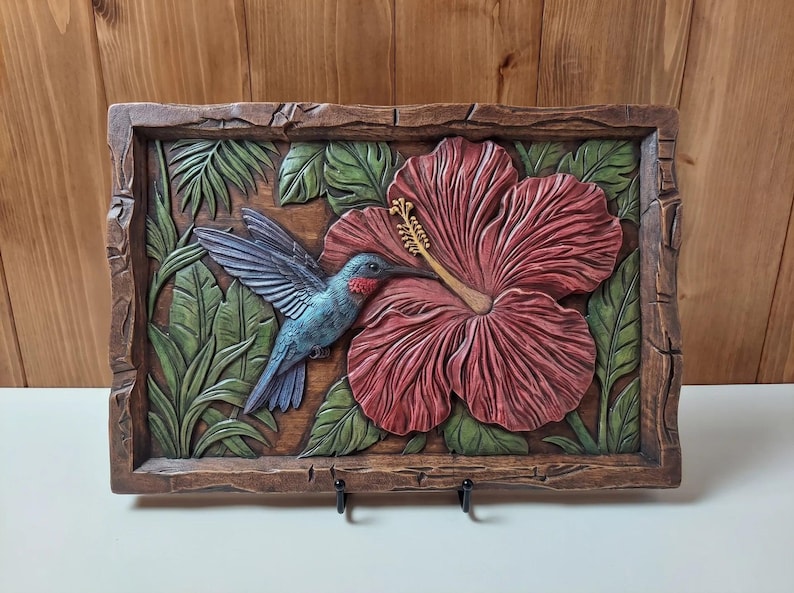 May include: A rectangular wooden wall plaque featuring a relief carving of a blue hummingbird with a red throat hovering near a large, textured red hibiscus flower. The scene is surrounded by green tropical leaves and a rustic, dark brown frame.