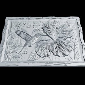 May include: A rectangular white wall plaque featuring a detailed relief carving of a hummingbird hovering near a large hibiscus flower. The design includes textured foliage in the background and a decorative border with a rough, stone-like edge.