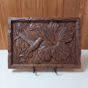 May include: A rectangular wooden wall plaque featuring a detailed relief carving of a hummingbird hovering near a large hibiscus flower, surrounded by tropical foliage. The piece has a dark brown finish and a textured, uneven border frame.