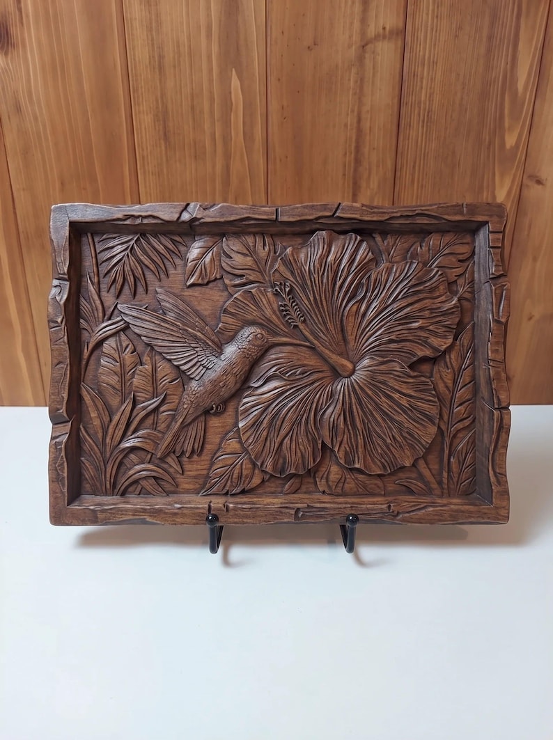 May include: A rectangular wooden wall plaque featuring a detailed relief carving of a hummingbird hovering near a large hibiscus flower, surrounded by tropical foliage. The piece has a dark brown finish and a textured, uneven border frame.