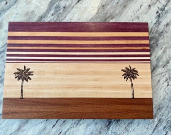 May include: A wooden cutting board with a striped design in shades of purple, brown, and beige. The board has two palm trees burned into the surface.