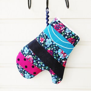 Puede incluir: Un guante de cocina colorido con un estampado floral, una sección rosa con lunares y rayas negras. El guante está colgado de un gancho de metal negro en una pared blanca. El guante tiene un lazo de cinta azul.