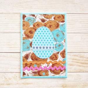 以下が含まれることがあります： イースターをテーマにした布製カード。水色の卵に青い水玉模様、茶色とターコイズのフローラルパターンが背景。カードは水色の縁取りと、オレンジとピンクの装飾トリムが施されています。