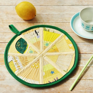 Puede incluir: Un salvamanteles circular con un borde verde y un diseño de rodaja de limón. El salvamanteles está hecho de tela amarilla y crema, con un acento de hoja verde. Un limón entero, una taza y un platillo, y una cuchara verde también están sobre la superficie de madera.