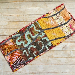 May include: A colourful patchwork table runner with a geometric design. The runner is made of different fabrics with animal prints and abstract patterns. It has a black and white trim.