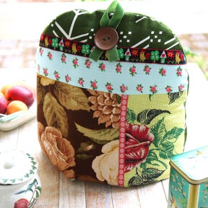 Peut inclure: Une housse de théière en tissu matelassé avec un motif patchwork comprenant une section supérieure vert foncé, une bande à fleurs bleu clair et une partie inférieure ornée de grandes roses brunes et jaunes. Un bouton brun et un ruban vert.