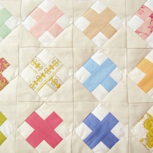 May include: Close-up of a handmade quilt with a repeating pattern of intersecting bands. The quilt features a cream-coloured background with colourful cross-shaped designs in shades of green, pink, blue, and yellow. The quilt is made of fabric.