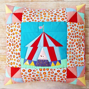 May include: Square cushion with a patchwork design. The centre features a circus tent with red and white stripes, set against a turquoise background. The surrounding patchwork includes floral and geometric patterns in various colours.