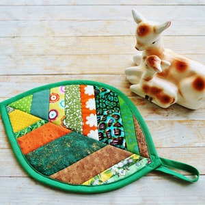 Peut inclure: Un dessous de plat en forme de feuille verte avec un motif patchwork de différentes couleurs et motifs, notamment vert, orange, jaune et marron. Le dessous de plat a une poignée en boucle.