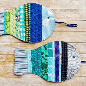 Könnte beinhalten: Zwei blaue und grüne Patchwork-Topflappen in Fischform mit einem schwarz-weißen Streifen am Schwanz. Die Topflappen haben ein blaues Band, das oben befestigt ist.