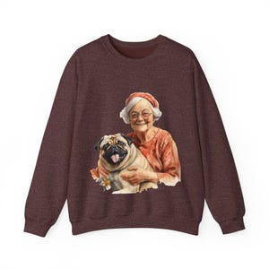 May include: Burgundy sweatshirt featuring a watercolor illustration of a smiling elderly person with glasses and a pug dog. The person is wearing an orange top and a matching hat. The pug has its tongue out.