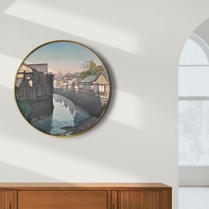 May include: A round wall art print featuring a Japanese woodblock print of a canal with buildings on either side. The print is in shades of gray, blue, and white.