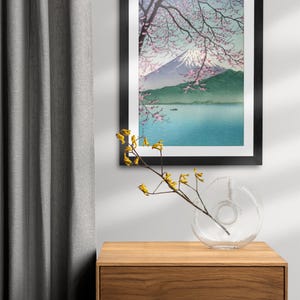 May include: A framed print of a Japanese landscape with Mount Fuji in the background, a lake in the foreground, and a cherry blossom tree in the foreground. The print is in a black frame and is hanging on a white wall.