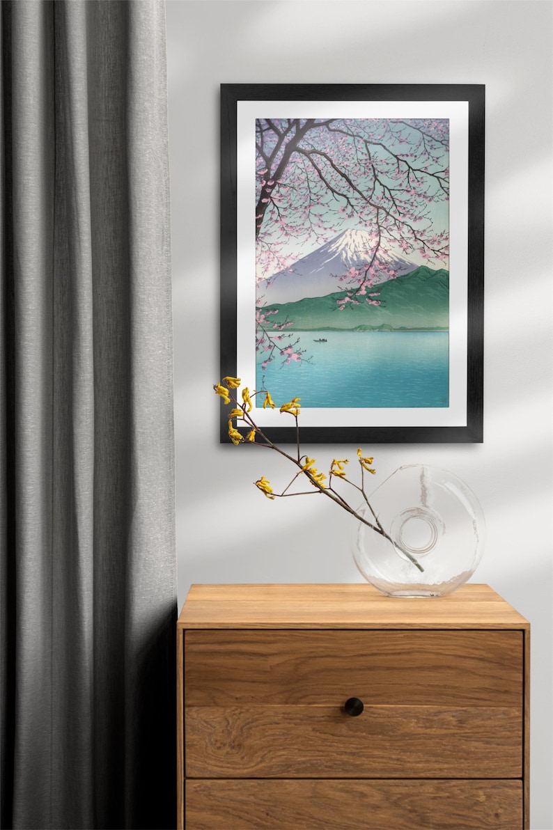 May include: A framed print of a Japanese landscape with Mount Fuji in the background, a lake in the foreground, and a cherry blossom tree in the foreground. The print is in a black frame and is hanging on a white wall.