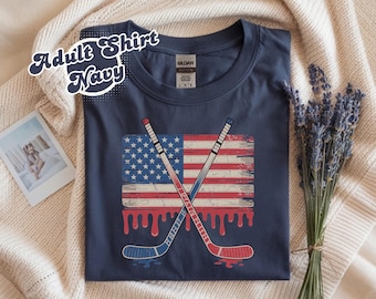 May include: Navy blue adult shirt featuring a distressed American flag design with crossed hockey sticks. The flag has red and white stripes, blue stars, and dripping red accents. The text "Adult Shirt Navy" is in a white, stylized font.