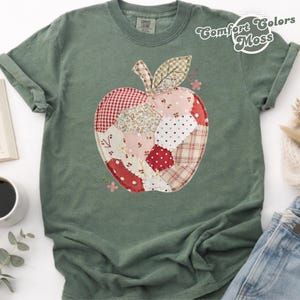 Op de afbeelding: Een mosgroen T-shirt met korte mouwen en een grote centrale appelafbeelding, opgebouwd uit diverse patchwork-stofpatronen zoals rood gingham, witte stippen, bloemenprints en ruitjes, voorzien van een klein bruin steeltje en een blaadje.