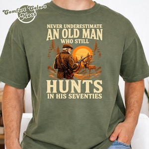 Op de afbeelding: Een olijfgroen t-shirt met een grafische print van een jager met een geweer en een hert in een bosrijke omgeving bij zonsondergang. De tekst luidt: Never underestimate an old man who still hunts in his seventies. Comfort Colors Moss logo is zichtbaar.